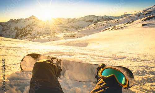 Snowboarder sitting on relax moment at sunset in french alps ski resort - Winter sport concept with person on top of the mountain ready to ride down - Legs view point with warm backlighting filter