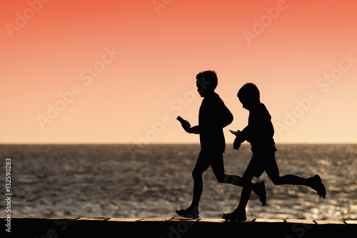 Silhouette of two boys running along the shore of the ocean with mobile phone in hand