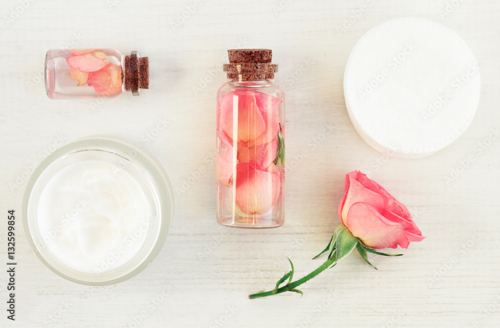Set of aromatic botanical cosmetic products on white wooden table. Glass bottles with extract, tonic infused with flower petals, fresh pink rose, cotton pads. Soft light and focus. Set of aromatic botanical cosmetic products on white wooden table. Glass bottles with extract, tonic infused with flower petals, fresh pink rose, cotton pads. Soft light and focus.