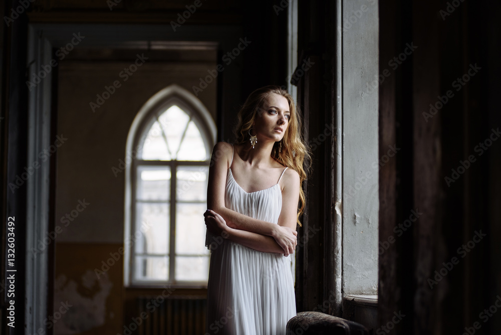 Naklejka premium Indoor summer portrait of young pretty cute girl. Beautiful woman posing beside fairytale window inside wood cabinet, scars old castle. Bride in white atlas dress. Photo has a model.