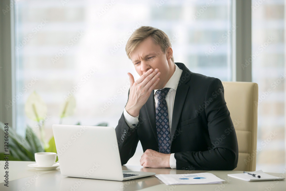 Young yawning businessman working with laptop at desk in the modern office, unmotivated employee, not completing job duties, do not have the drive to succeed, dissatisfaction in office environment Young yawning businessman working with laptop at desk in the modern office, unmotivated employee, not completing job duties, do not have the drive to succeed, dissatisfaction in office environment