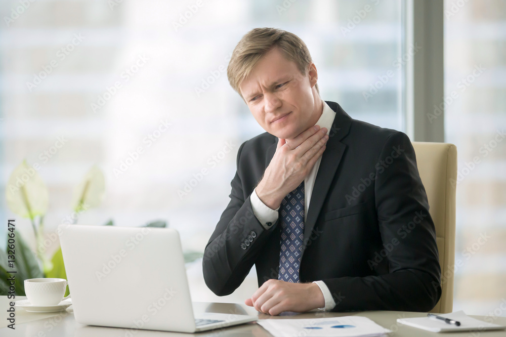 Young businessman working with laptop at desk in the office, hand at his neck, feeling unwell, have a sore throat, after loud screaming, loss of voice, irritation, pain, and itchiness, hard to swallow Young businessman working with laptop at desk in the office, hand at his neck, feeling unwell, have a sore throat, after loud screaming, loss of voice, irritation, pain, and itchiness, hard to swallow