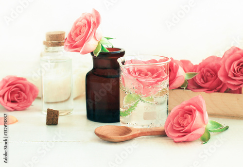 Rose cosmetic ingredients for herbal spa beauty treatment. Flower in attar glass test jar with ml scale on wooden white table. Soft focus, soft light.