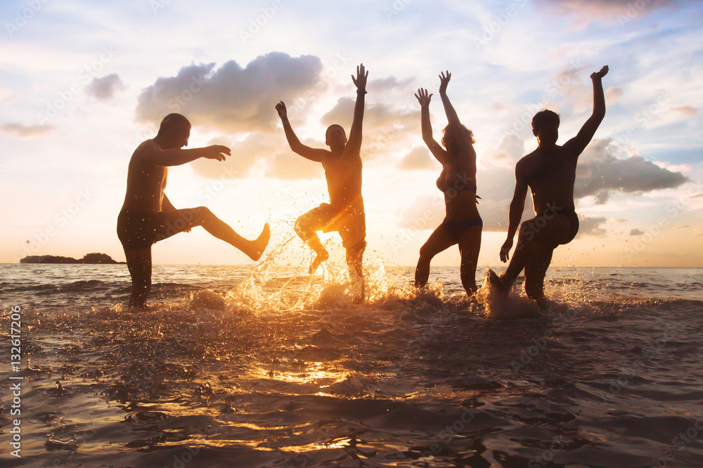 group of happy friends having fun together on the beach at sunset, jumping and dancing with water splash in the sea, silhouettes of people