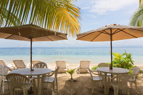 Fototapeta Naklejka Na Ścianę i Meble -  Restaurant tables, sun lounger and beach umbrella under the palm leaves on the beach