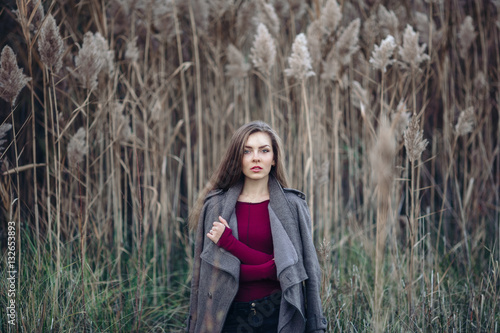 Portrait of pensive sad lonely Caucasian blonde young beautiful woman girl with long hair wearing jeans, coat jacket, in forest field among large tall plants grass looking in camera