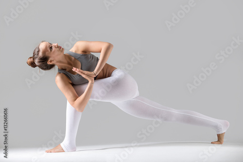 Young attractive woman practicing yoga, standing in Revolved Side Angle exercise, Parivrtta Parsvakonasana pose, namaste, working out wearing sportswear, indoor full length, grey studio background