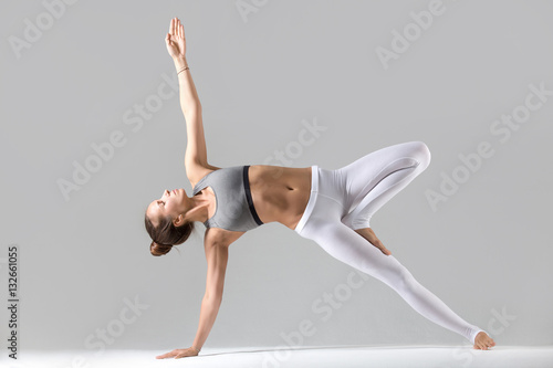 Young attractive woman practicing yoga, stretching in Side Plank exercise, Vasisthasana pose, working out wearing sportswear, white pants, indoor full length, isolated against grey studio background