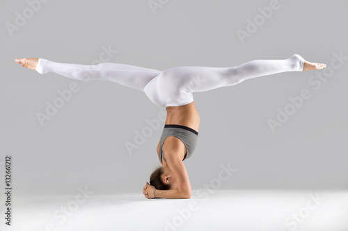 Young woman practicing yoga, standing in headstand exercise, salamba sirsasana pose, working out wearing sportswear, white pants, bra, indoor full length, isolated against grey studio background