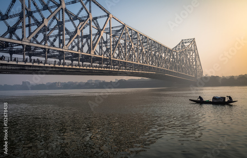 Historic Howrah bridge on Ganga river on a misty winter morning with a wooden fishing boat passing the bridge. Kolkata, India.