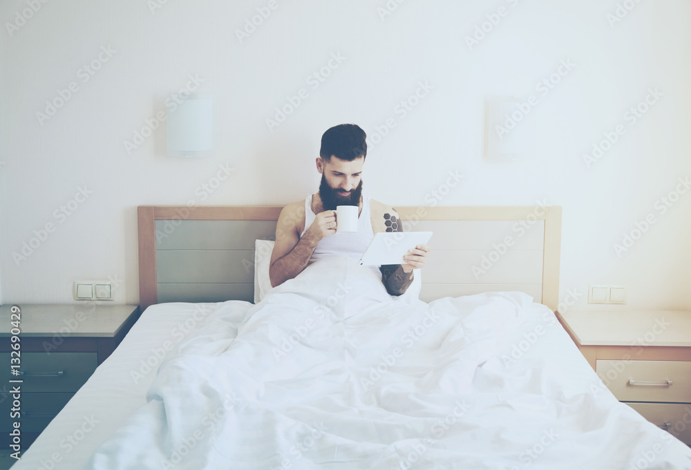 Fototapeta premium Bearded man lying in morning bed with coffee or tea cup and digi