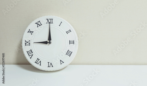 Closeup white clock for decorate show 9 o'clock on white wood desk and cream wallpaper textured background with copy space