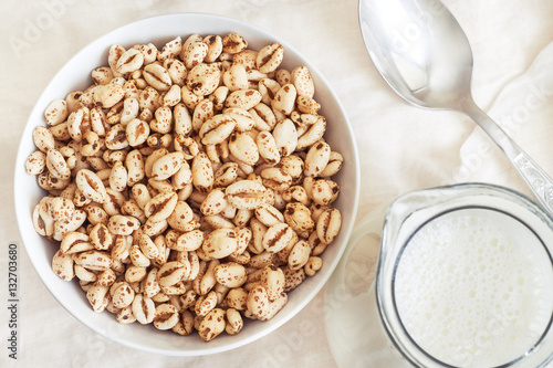 Puffed wheat cereal in white bowl with pitcher of milk and spoon. Top view