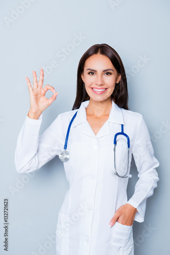 Young happy cheerful doctor in white uniform gesturing ok