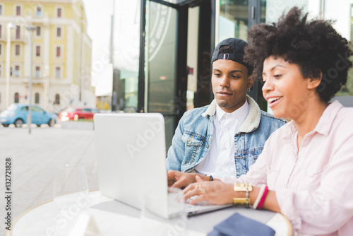 Young multiethnic couple using computer sitting outdoor in the city - business, technology, internet concept
