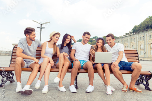Young cheerful friends enjoying weekend and sitting on bench wit