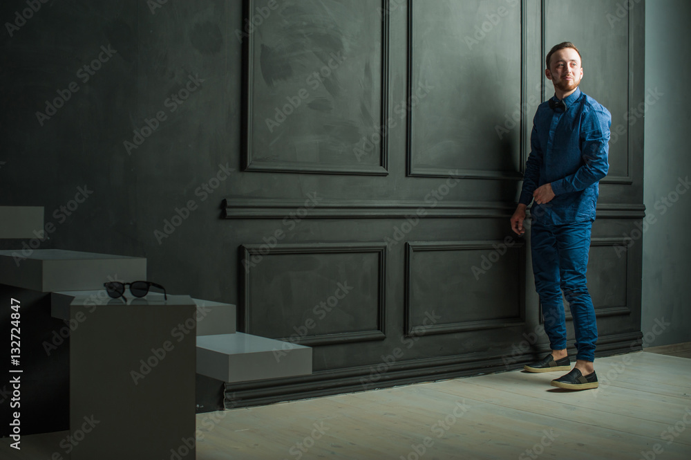 studio portrait of a young man on a dark wall background in a blue shirt and jeans. hipster man, dressed stylishly, with a beard studio portrait of a young man on a dark wall background in a blue shirt and jeans. hipster man, dressed stylishly, with a beard