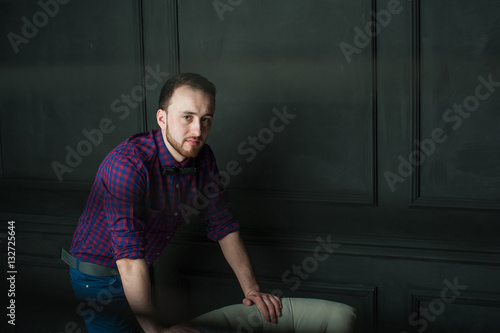 studio portrait of a young man on a dark wall background in a burgundy plaid shirt and blue jeans. hipster man, dressed stylishly, with a beard, in a room with an armchair