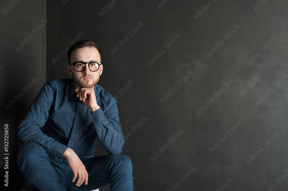 studio portrait of a young man on a dark wall background in a blue shirt and jeans. hipster man, dressed stylishly, with a beard and glasses, sitting on white stairs studio portrait of a young man on a dark wall background in a blue shirt and jeans. hipster man, dressed stylishly, with a beard and glasses, sitting on white stairs
