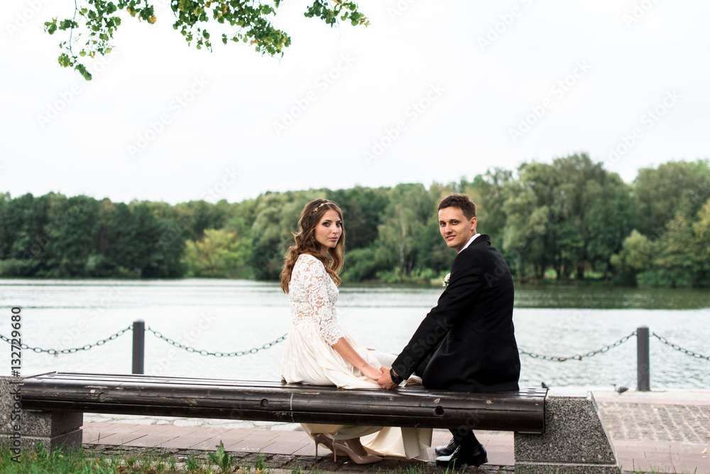 happy bride and groom at a park on their wedding day sitting on a bench near the lake