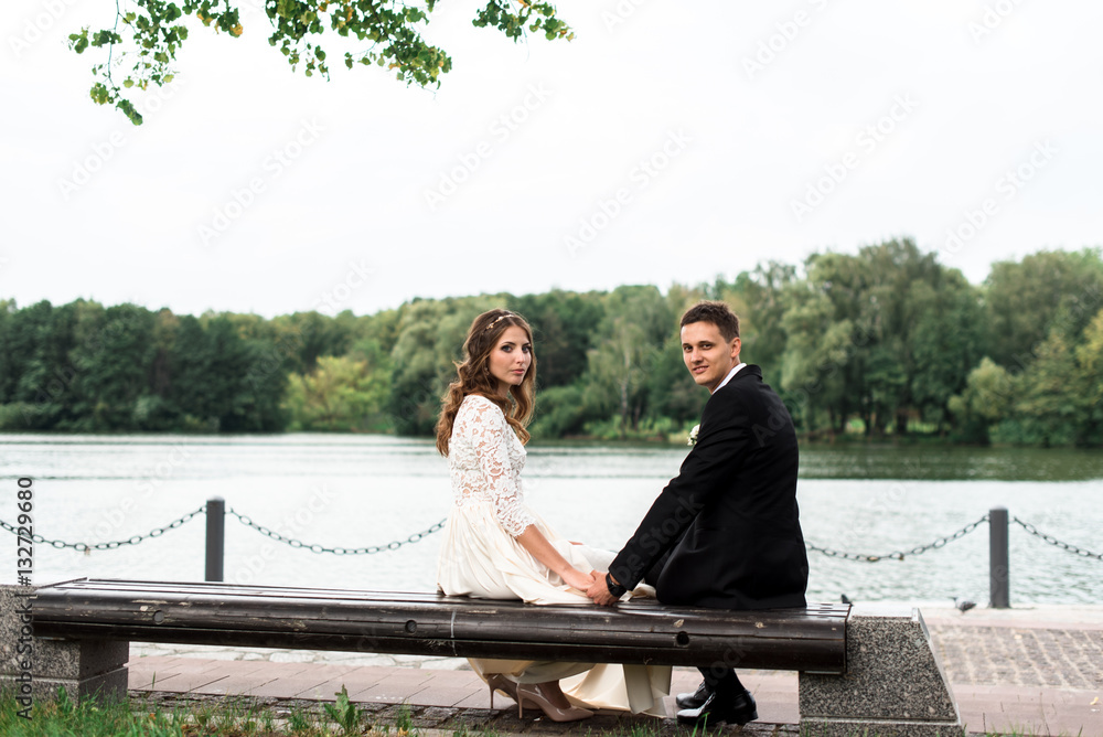 happy bride and groom at a park on their wedding day sitting on a bench near the lake happy bride and groom at a park on their wedding day sitting on a bench near the lake