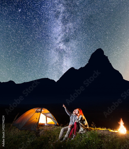 Romantic couple covered with a plaid is sitting near the glowing tent and campfire and looking to the stars and Milky way in the night sky. Silhouette of the mountains on the background. Long exposure
