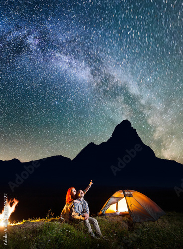 Tourist family - man and woman looking at the shines starry sky at night. Couple hikers sitting near camp and campfire. Milky way and mountains on the background