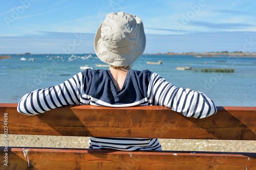 Une jeune femme habillée d'un pull marin est assise sur un banc face à la mer