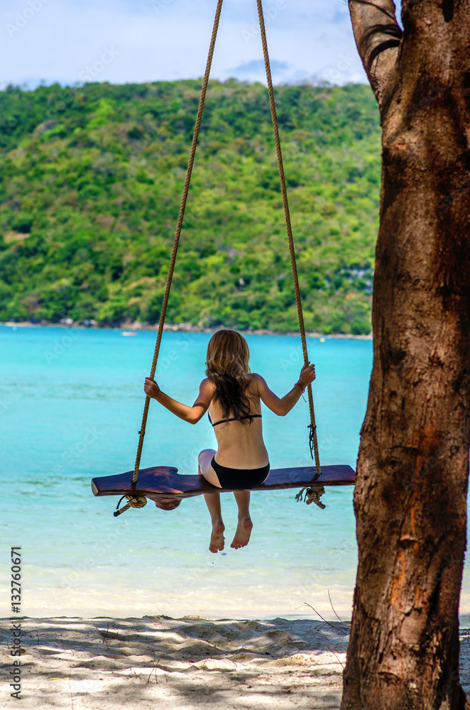 Happy girl swinging and relaxing on a rope swing with a paradise tropical sea in background - concept about summer vacations