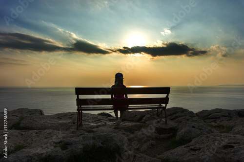 The silhouette of a lonely girl sitting on a bench on the top of the edge of a cliff and watching the sunset