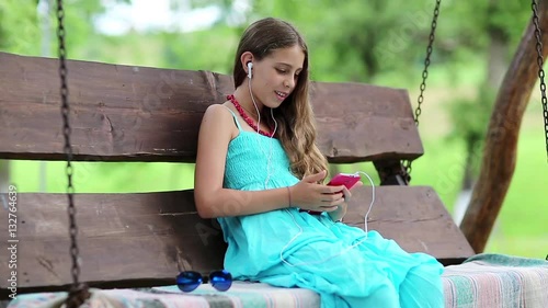 Beautiful girl in blue dress sits on the swing bench in garden and speaks via red smartphone. Attractive girl with earphones and smartphone communicates through skype. Female with smartphone photo