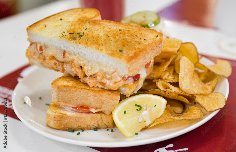 A cheese, prawn, seafood and tomato grilled sandwich, served with crisps and a wedge of lemon, on a white plate.