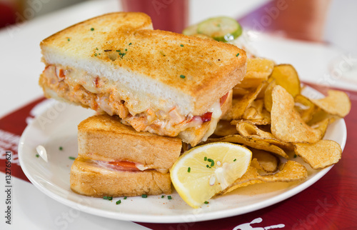 A cheese, prawn, seafood and tomato grilled sandwich, served with crisps and a wedge of lemon, on a white plate.