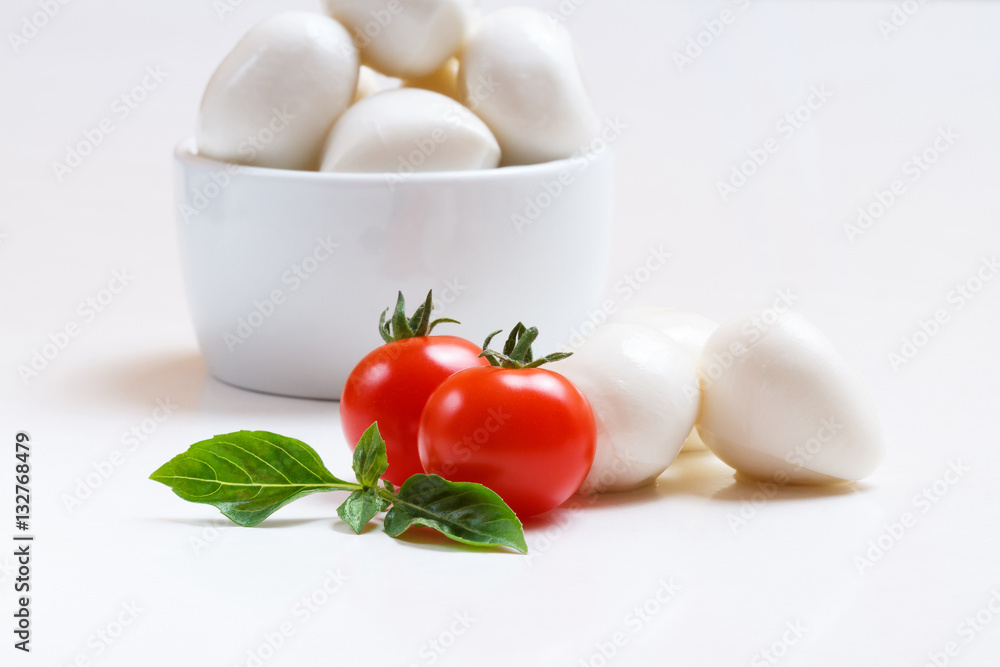 Small mozzarella balls in white bowl with cherry tomatoes and fresh basil on white background