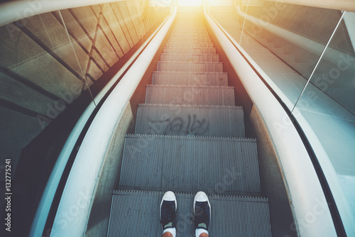 Outdoor city escalator stairway down, feet of guy in gumshoes ready to go down; evening sun, with tiled concrete wall on the left, wide view from above, vintage color filter