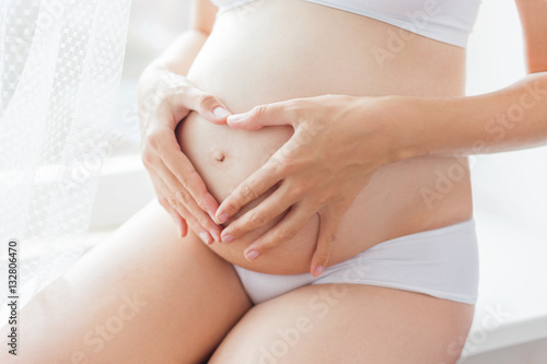 Pregnant woman in white underwear. Young woman expecting a baby. Future mother makes heart gesture - symbol of love.