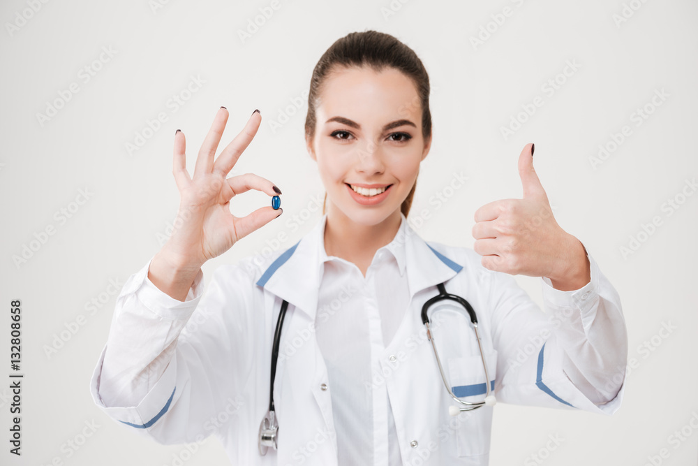 Cheerful woman doctor holding a pill and showing thumbs up