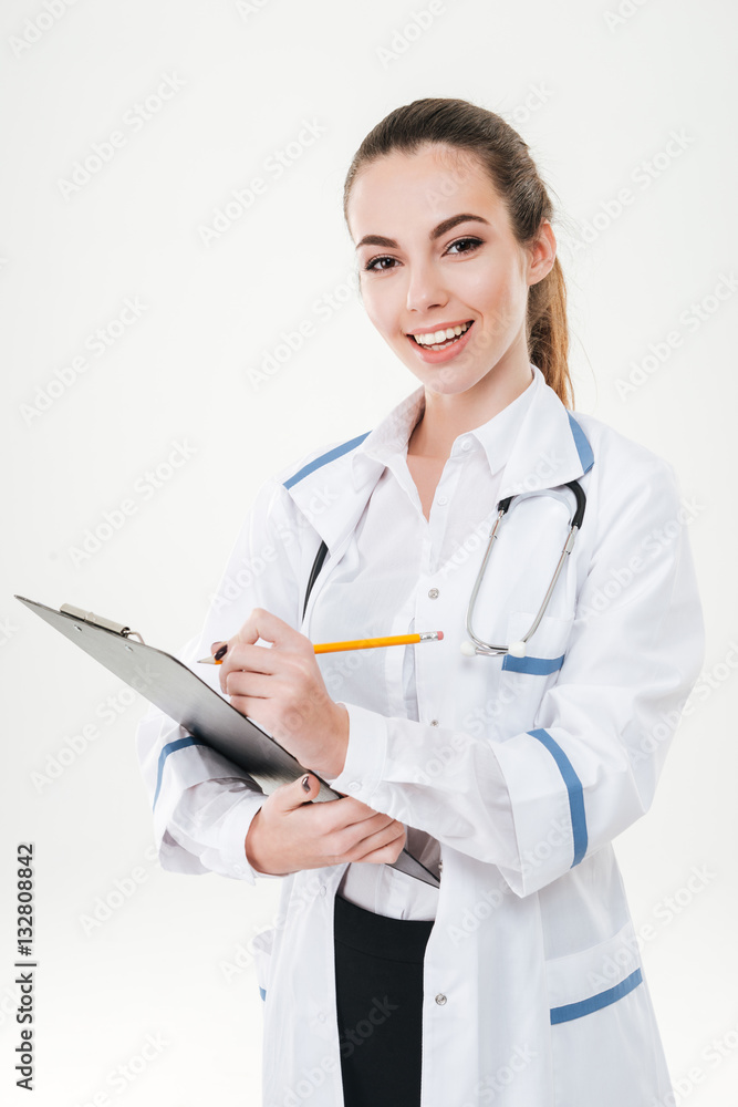 Cheerful cute young woman doctor standing and writing on clipboard Cheerful cute young woman doctor standing and writing on clipboard