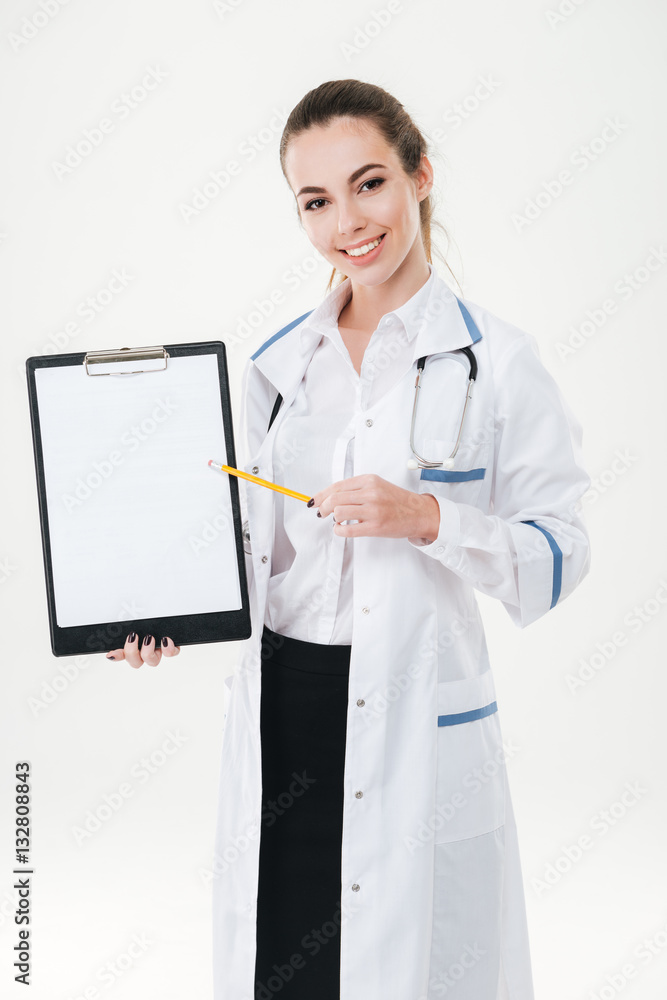Smiling young woman doctor standing and pointing on blank clipboard Smiling young woman doctor standing and pointing on blank clipboard