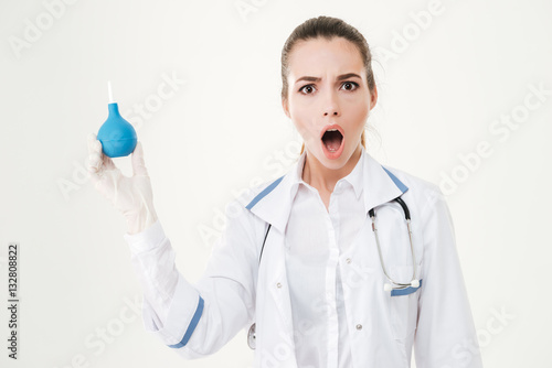 Shocked young woman doctor in gloves holding enema photo