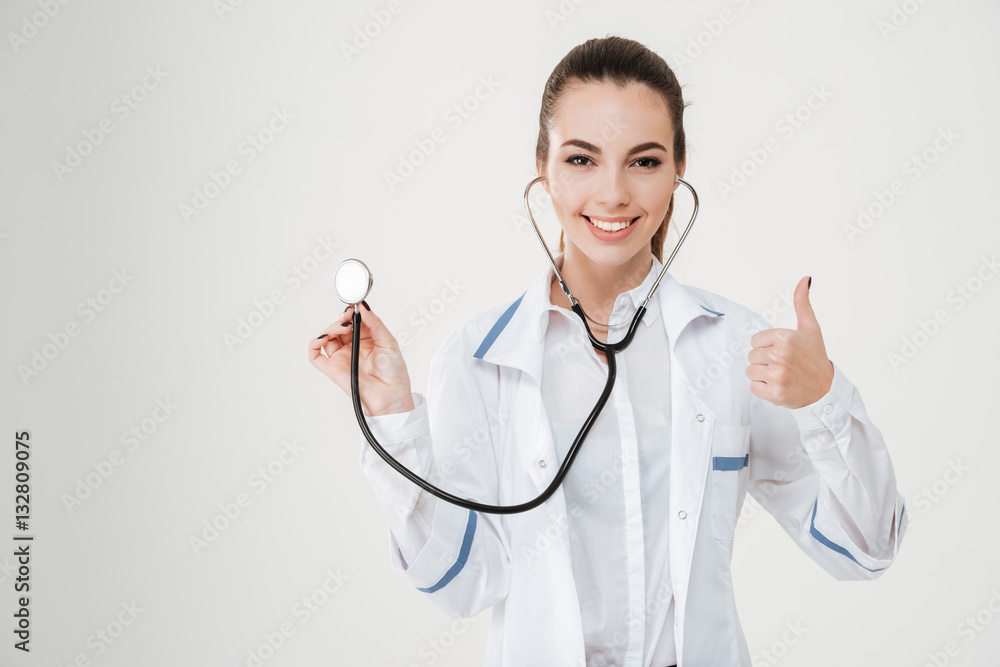 Cheerful young woman doctor using stethoscope and showing thumbs up Cheerful young woman doctor using stethoscope and showing thumbs up