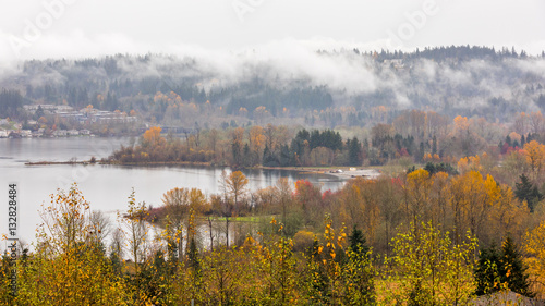 River bank in the mist, autumn forest on the riverside, beautiful trees and small houses on a background of mountains