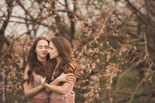 Beautiful happy young twins sisters in long evening dresses enjoying smell in a blooming spring garden. Having fun together  positive emotions  bright colors. Copy space. Healthy hair  clean skin.