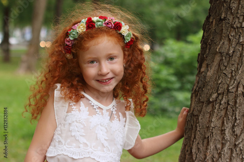 Little girl with flowers wreath poses near big tree in summer pa