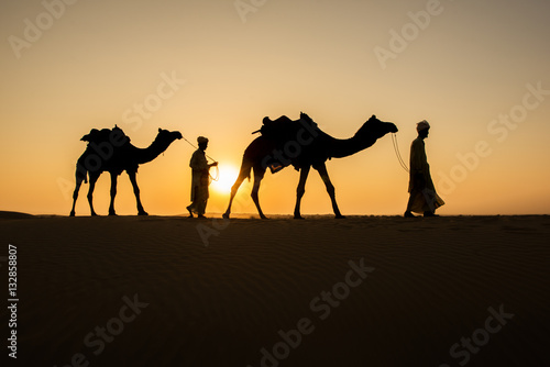 Rajasthan travel background - two indian cameleers  camel drivers  with camels silhouettes in dunes of Thar desert on sunset. Jaisalmer  Rajasthan  India