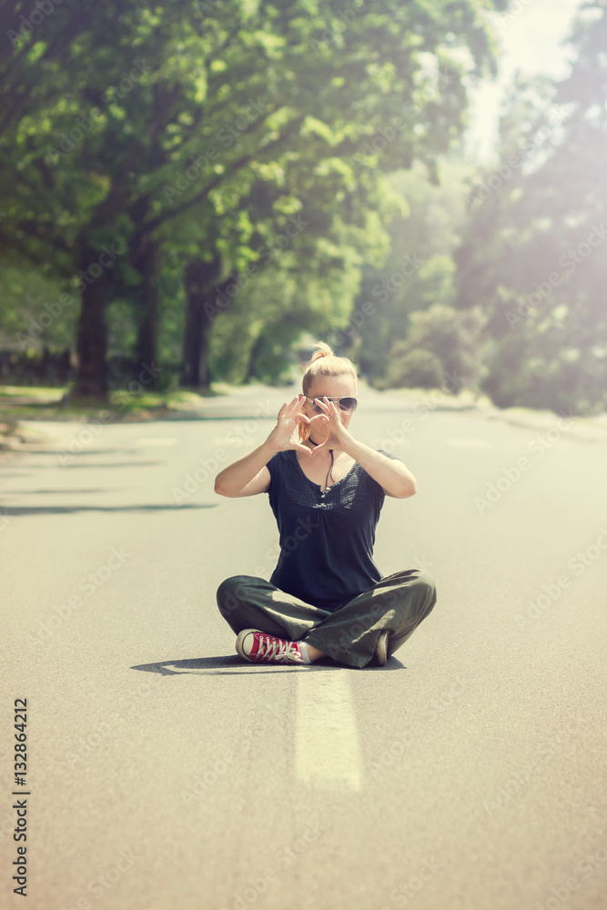Woman sitting with crossed legs in the middle of the street, making heart shape with her hands