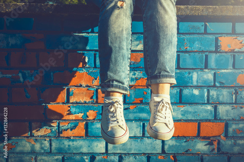 Billede på lærred Female legs in white canvas sneakers and jeans dangling from a blue brick wall o