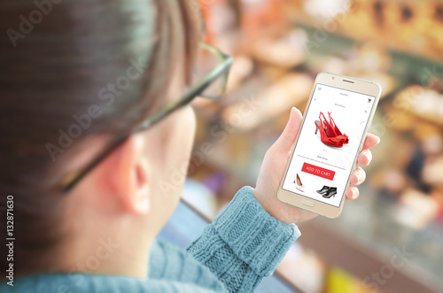 Woman shopping online with smart phone. Commerce web site or app on mobile display. Woman red shoes and add to cart button. Shopping center in background.