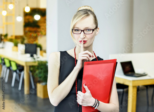 Beautiful blondie office with folders in hands puts her finger to her lips in an office.