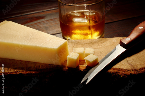 Parmesan cheese cut into cubic on a wooden board with knife and glass of whiskey on the second plan with ice